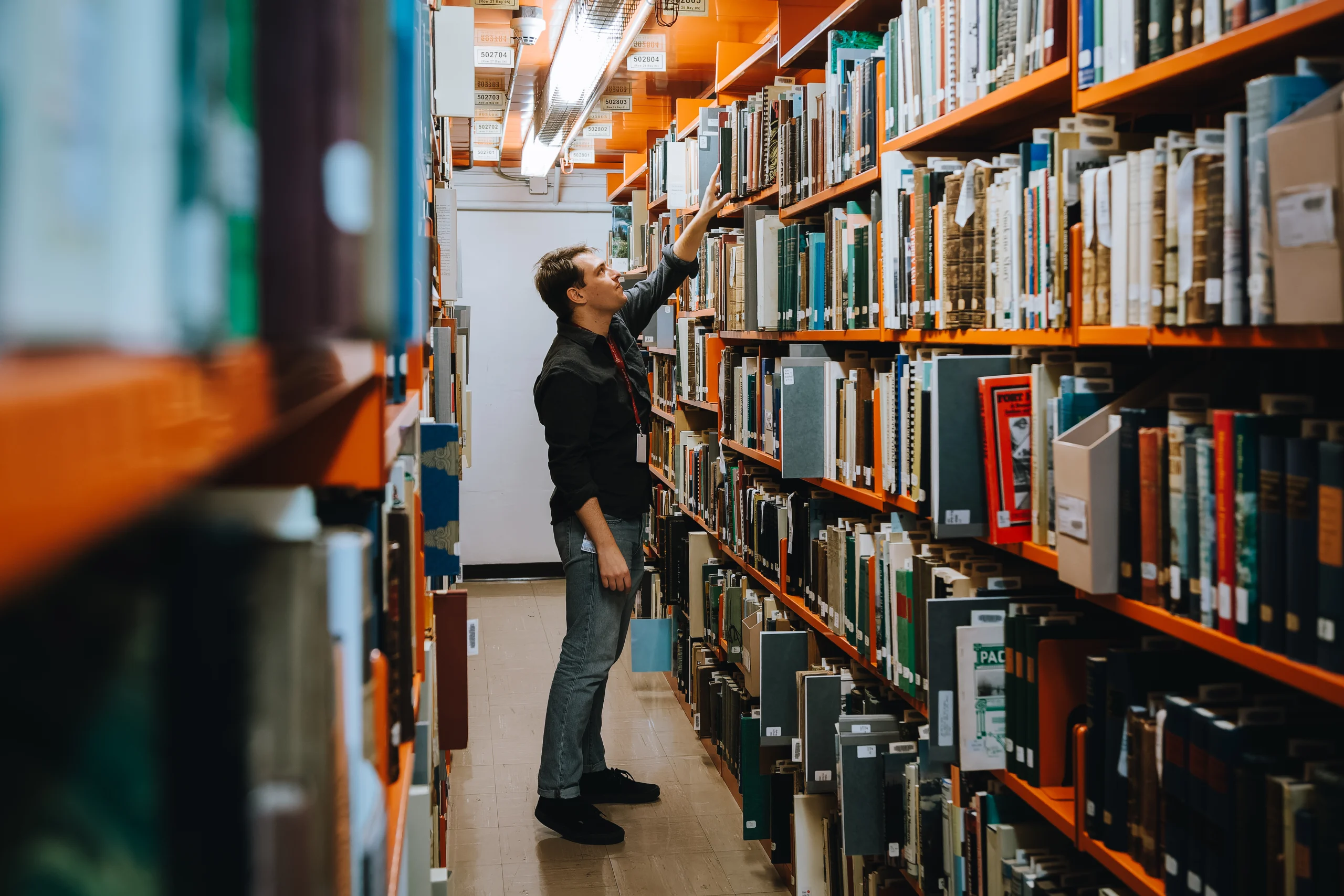 A person with short hair and a black shirt reaches for a book on the highest shelf of a packed archival bookshelf.