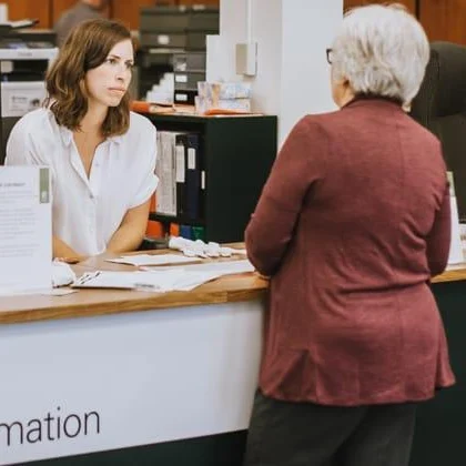 A woman with short white hair in a red jacket speaks with a woman wearing a white shirt sitting behind an information desk.